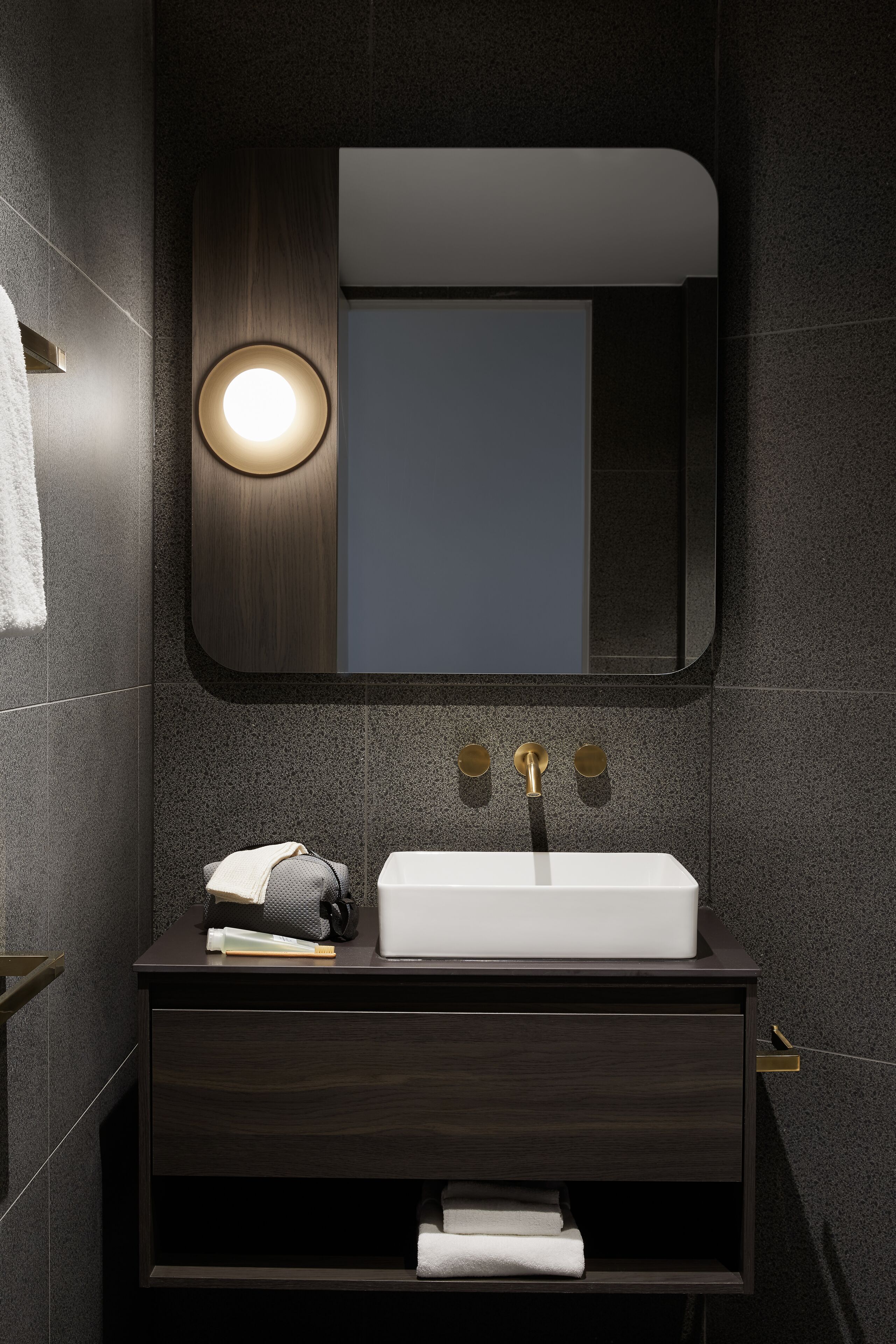 Un baño moderno con tocador de madera oscura, un lavamanos blanco y un espejo redondo con luz superior.