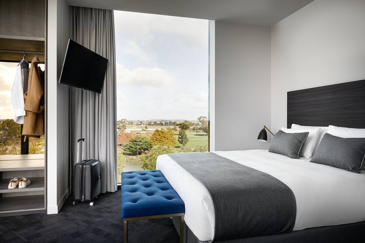 A modern hotel room with a large bed, a flat-screen TV, a view of the outdoors, and a seating area with a suitcase.