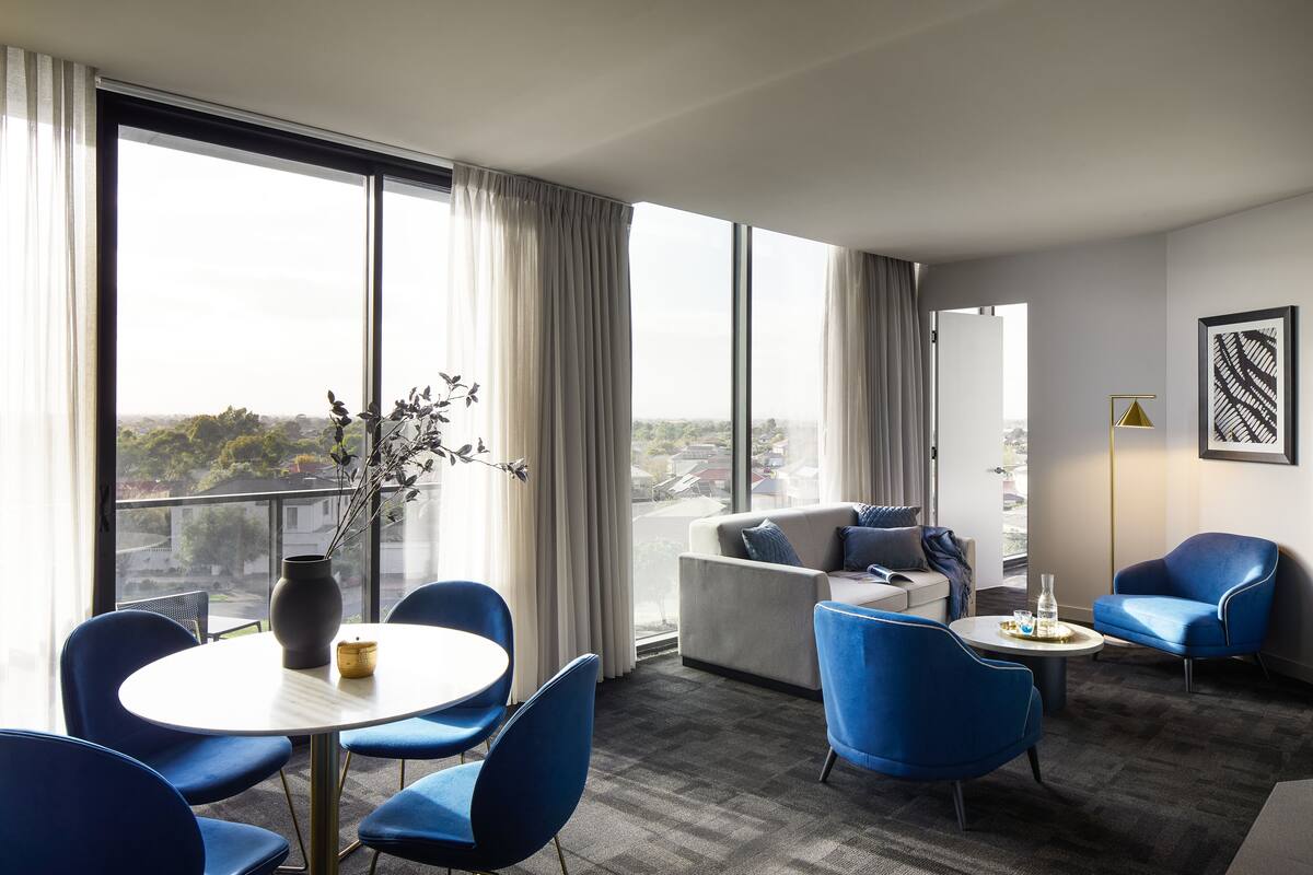 A modern hotel room with a large window, a round table with chairs, a sofa, and a blue armchair.