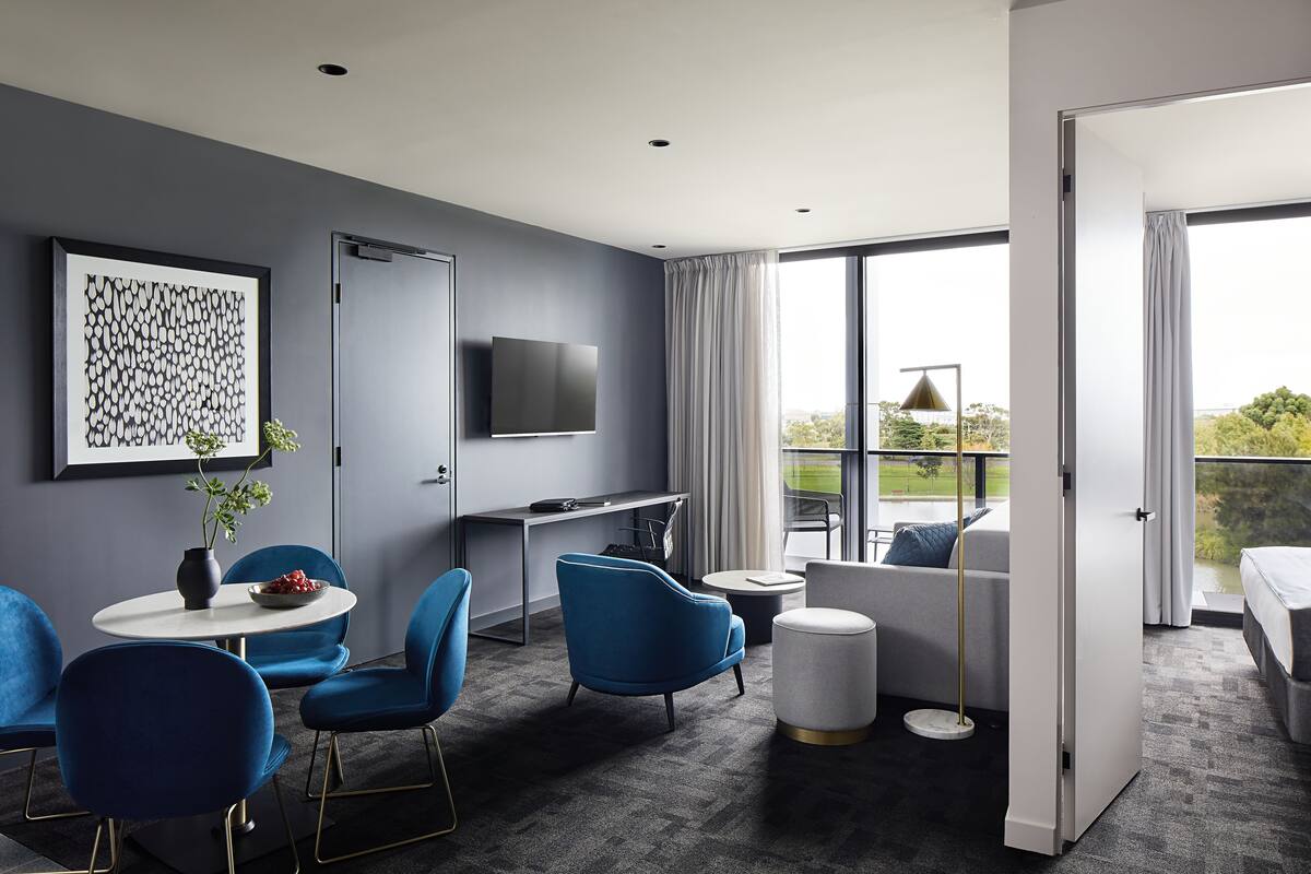 A modern hotel room with a grey wall, a flat-screen TV, a dining area with blue chairs, a round table, and a large window with curtains.
