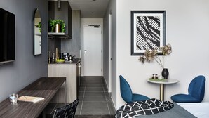 Habitación de hotel moderna con un escritorio de madera oscura, una pequeña mesa redonda con un florero y una taza, dos sillas azules, una cama con una colcha de diseño blanco y negro, un espejo grande y una obra de arte abstracta enmarcada en la pared.