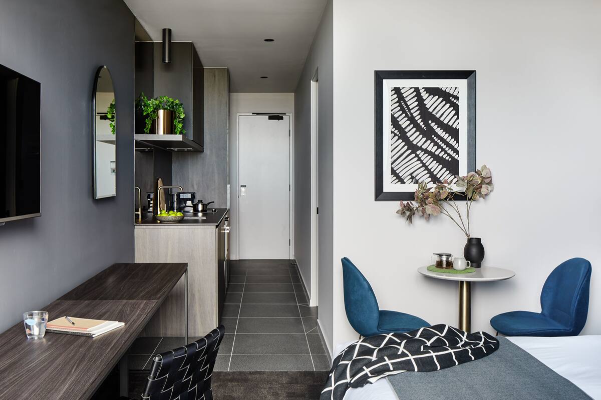 A modern hotel room with a dark wood desk, a small round table with a vase and a cup, two blue chairs, a bed with a black and white patterned blanket, a large mirror, and a framed abstract artwork on the wall.