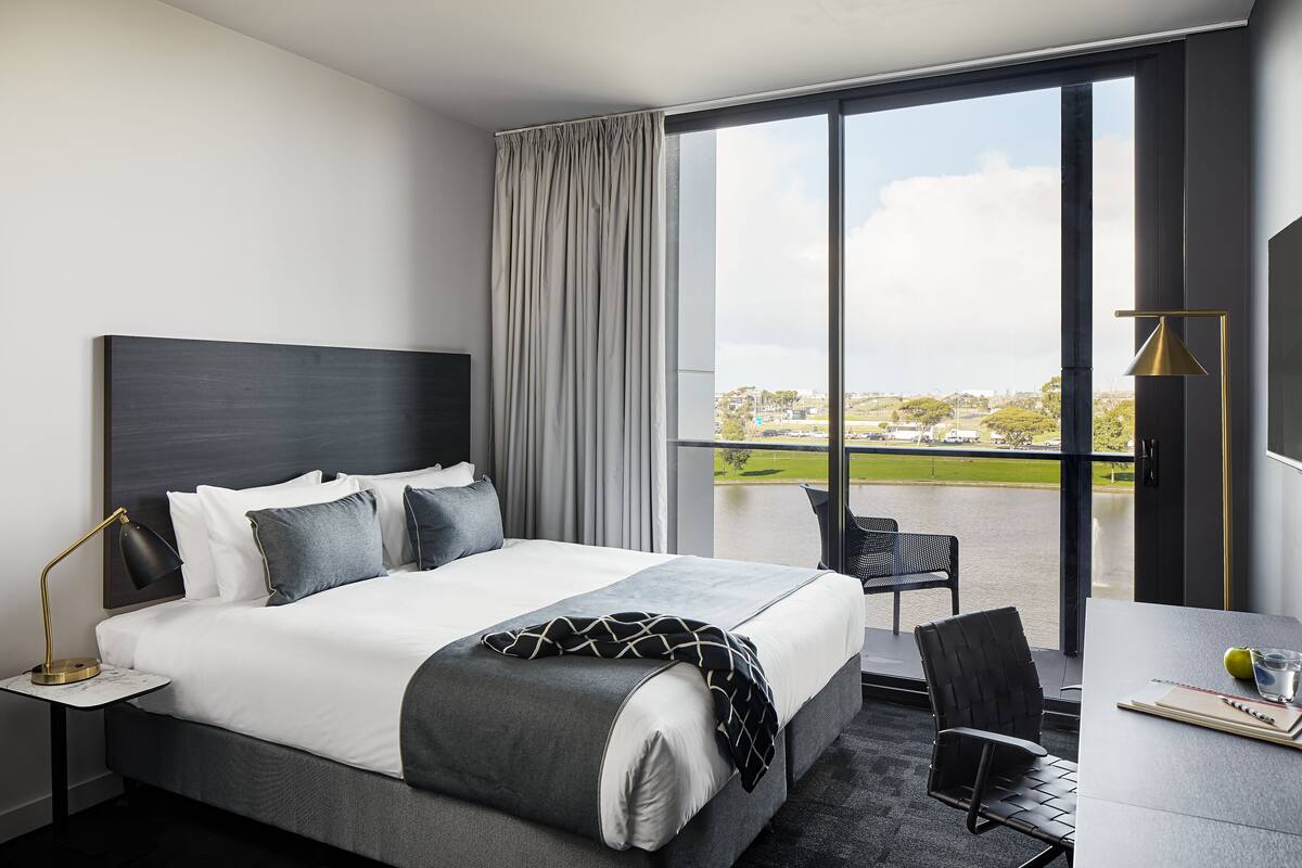 A modern hotel room with a large bed, a desk with a chair, and a view of the city through a large window.