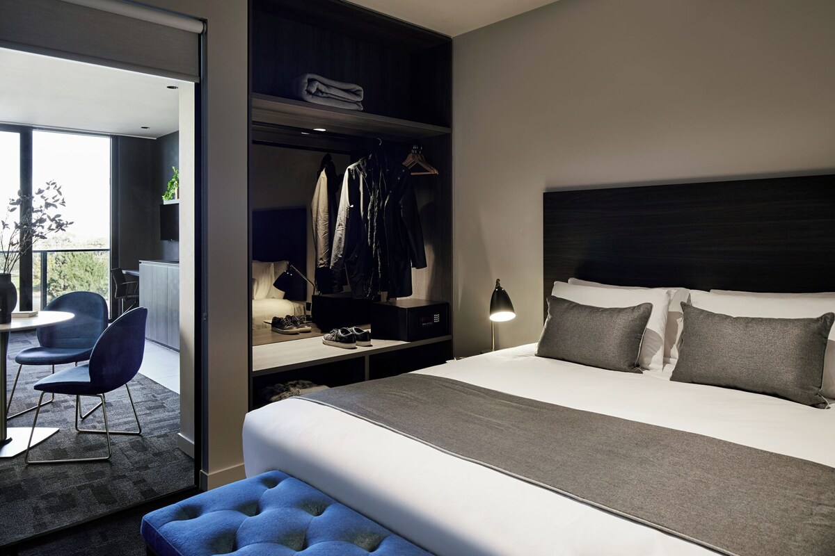 A modern hotel room with a large bed, a desk, a chair, and a walk-in closet.