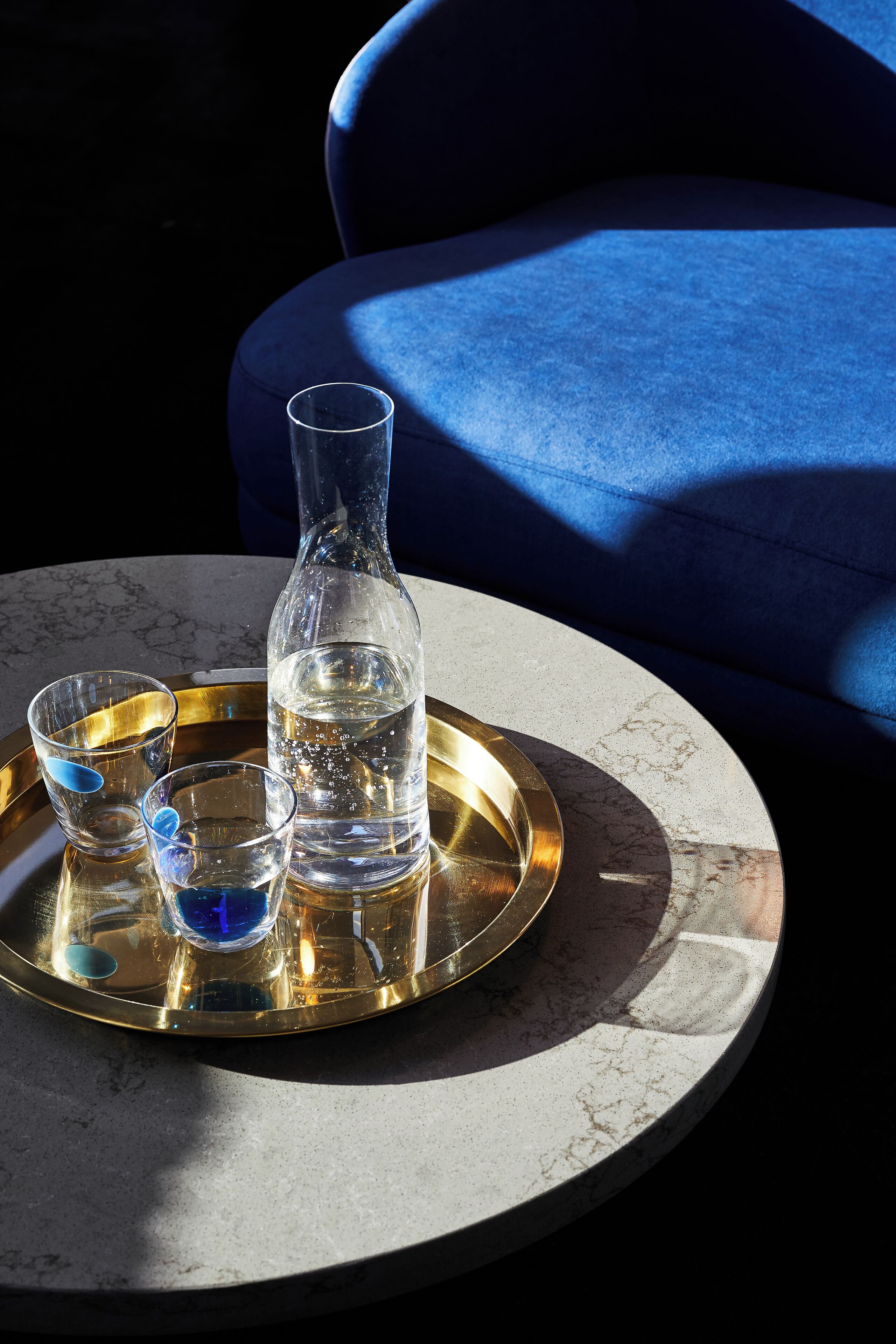 Une table ronde surmontée d’un plateau en marbre, un plateau doré, une carafe en verre transparent et deux verres à liqueur remplis d’un liquide bleu.