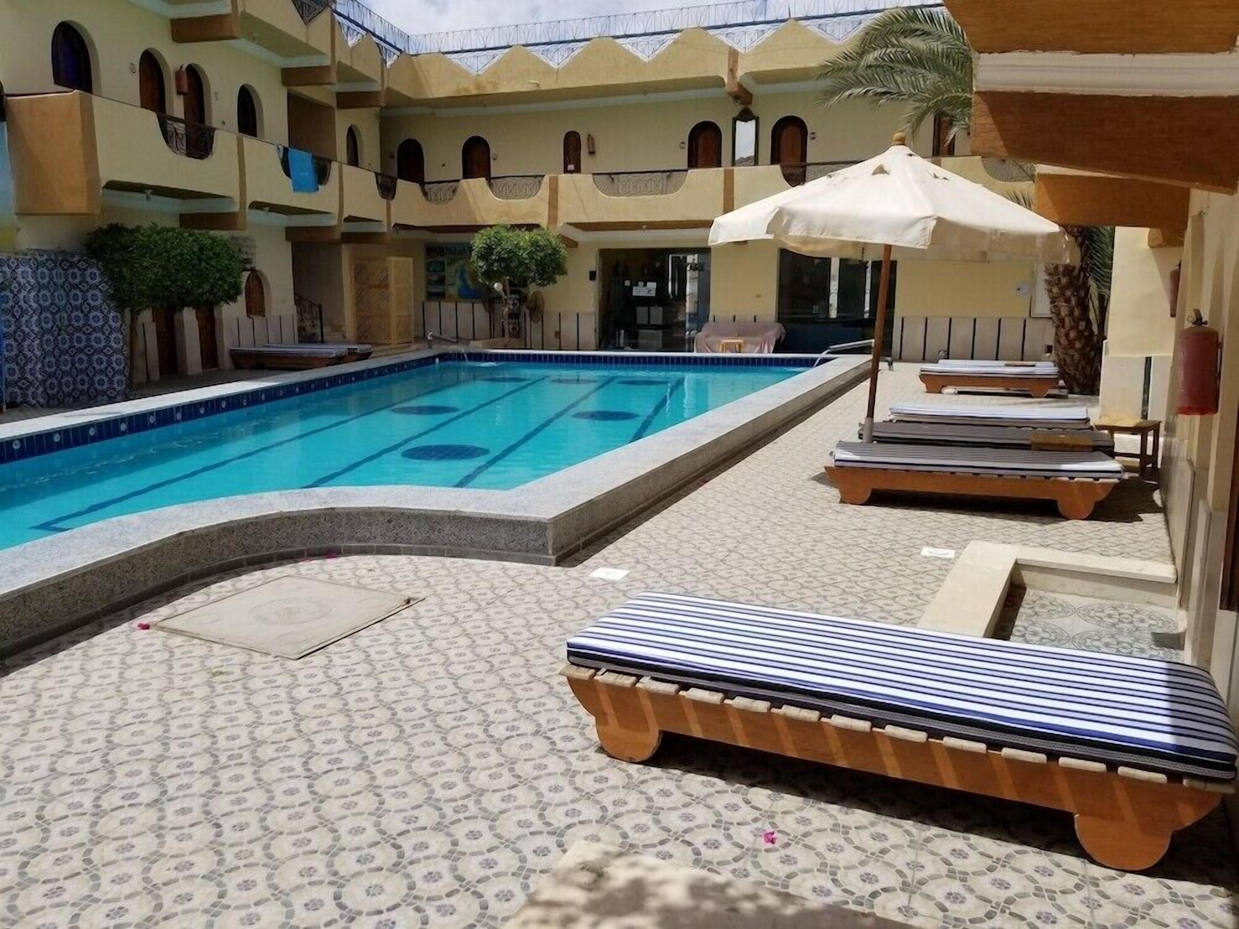A swimming pool with lounge chairs and umbrellas, surrounded by a patterned tiled floor and a building with arches and balconies.