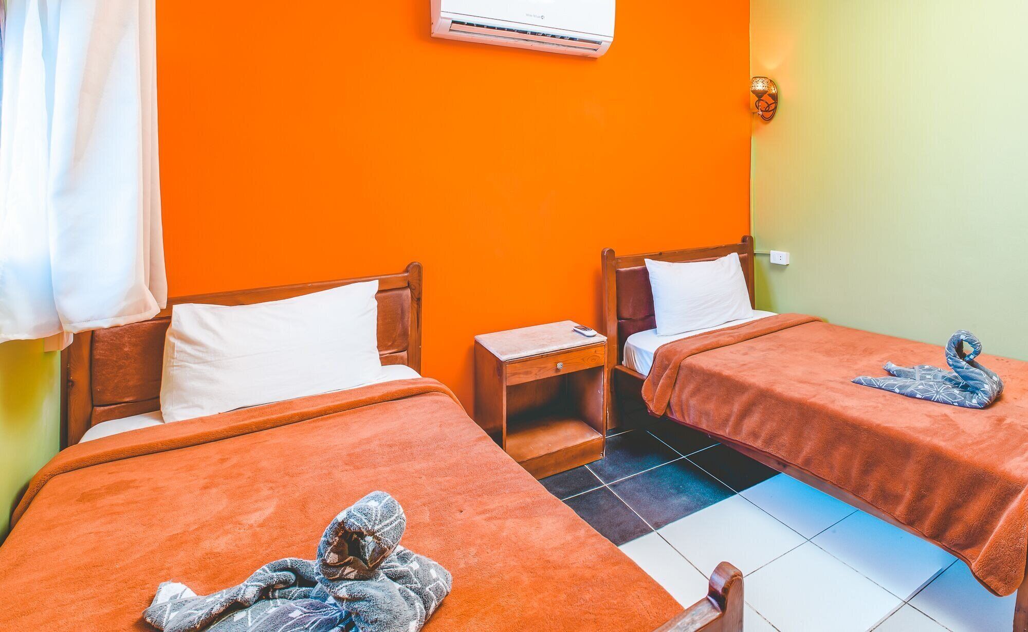 A room with two beds, each with a wooden headboard and white pillows. There is a small wooden nightstand with a remote control on it. The walls are orange, and there is an air conditioning unit mounted on the wall.