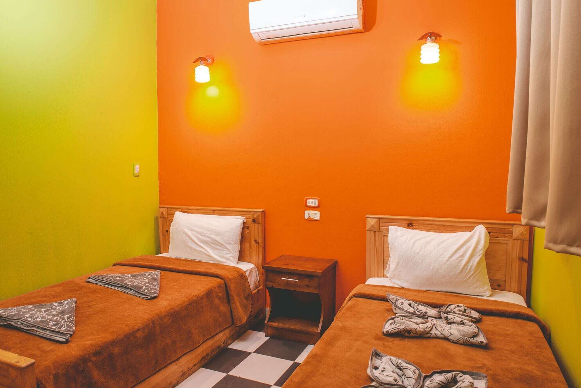 A room with two beds, each with a wooden headboard and a white pillow. There is a small wooden nightstand between the beds. The walls are painted orange, and there is a window with curtains.