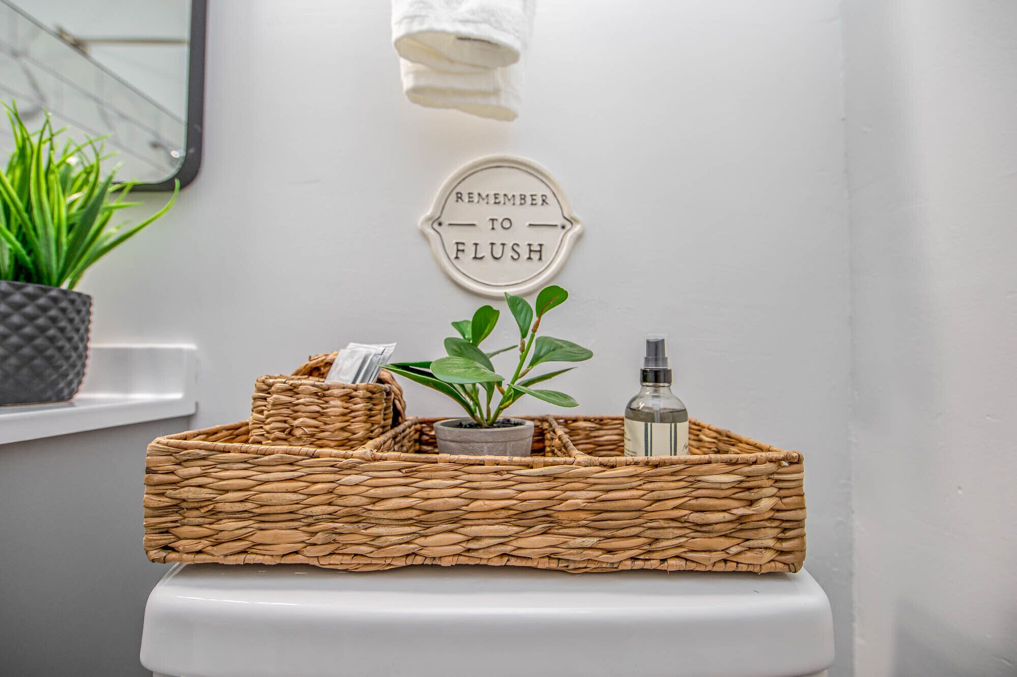 A bathroom with a toilet, a wicker basket on the toilet, a potted plant, a bottle, and a sign that says "REMEMBER TO FLUSH".