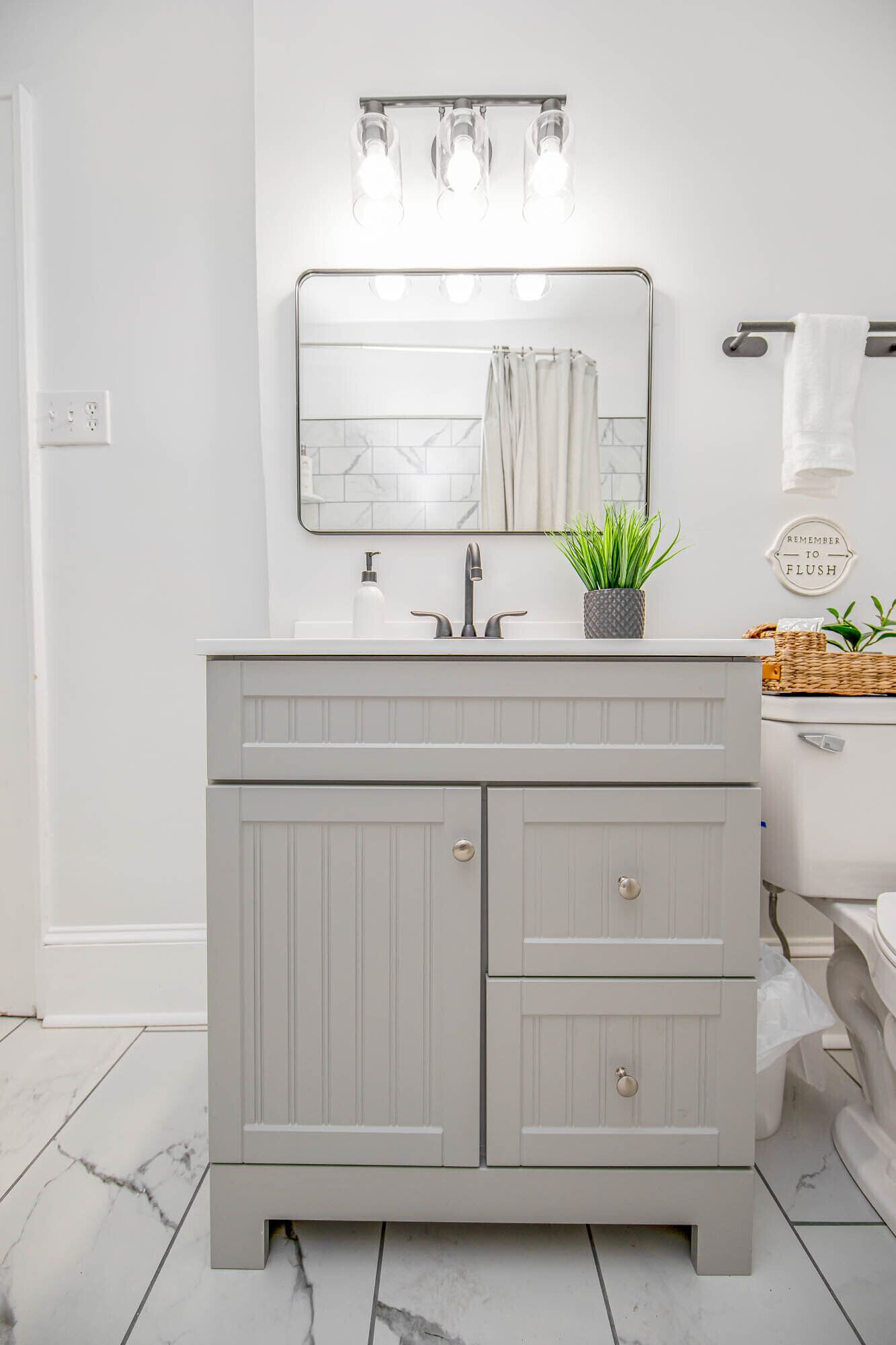 A bathroom with a white vanity, a mirror, a light fixture above the mirror, a potted plant on the vanity, and a sign on the wall.