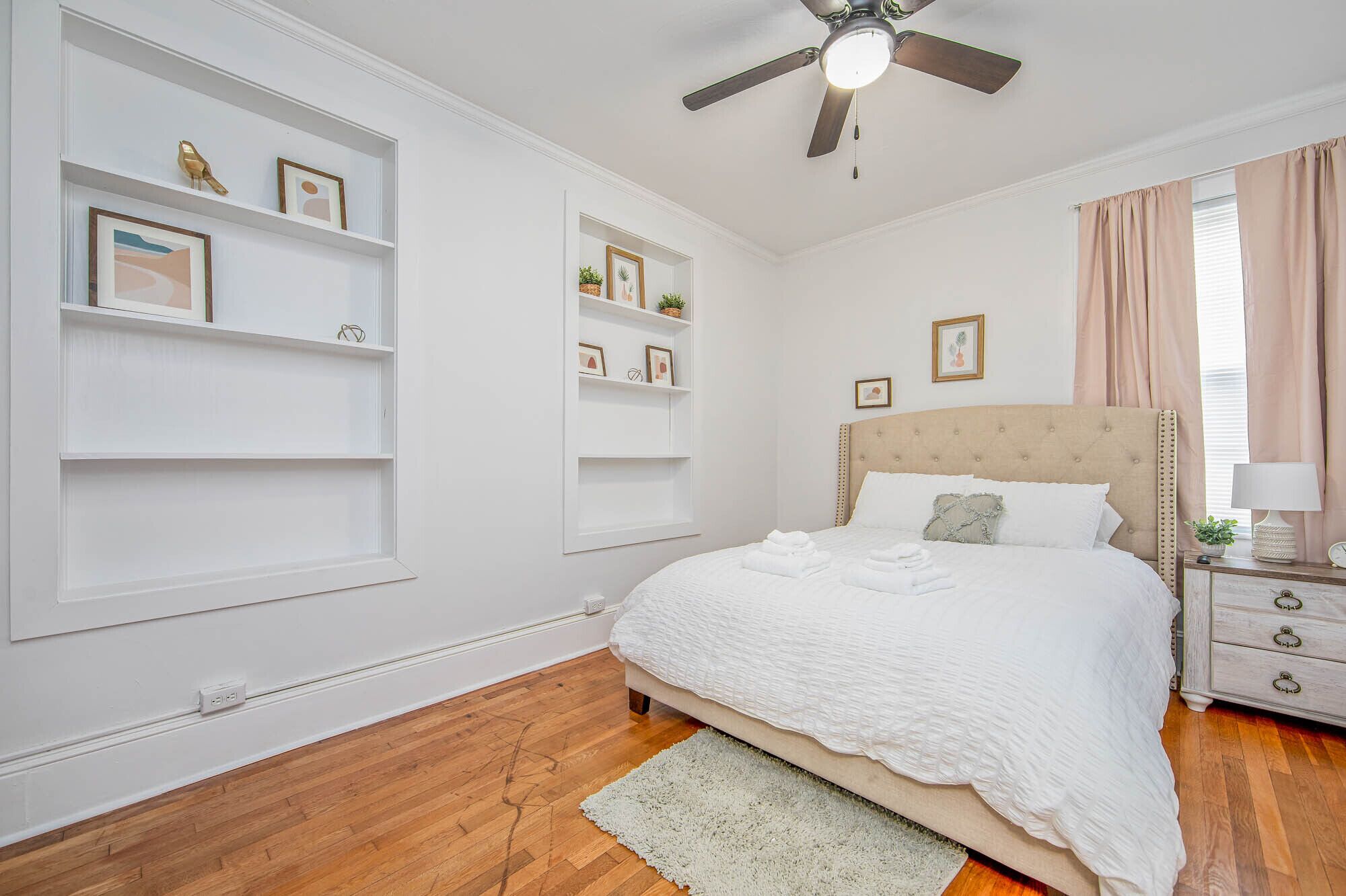 A bedroom with a bed, bedside table, lamp, and wall-mounted shelves with framed pictures.