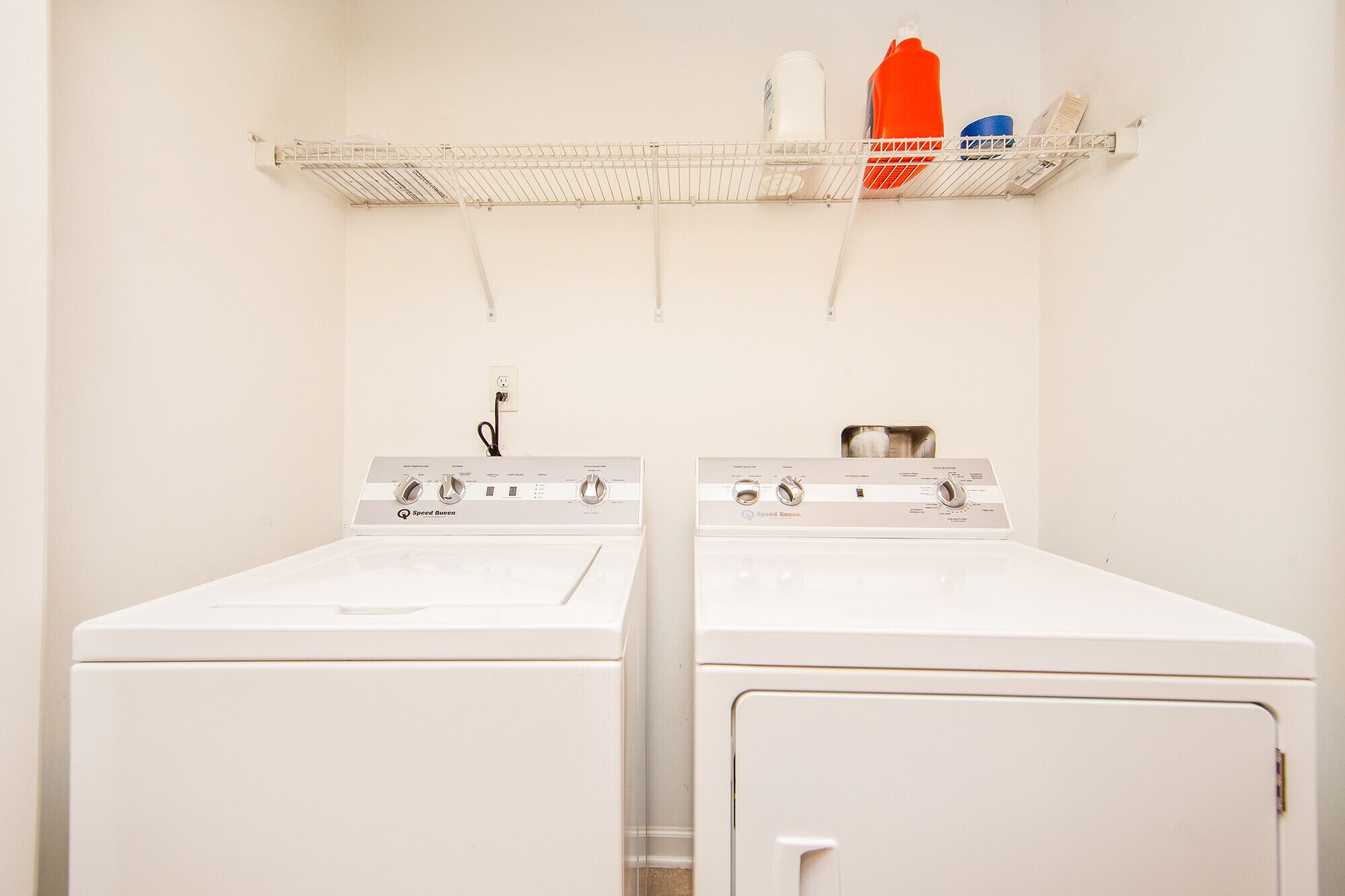 A laundry room with two white Speed Queen washers and dryers, a metal shelf above holding cleaning supplies, and a white wall.