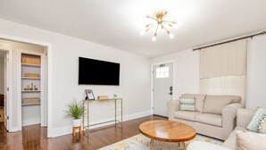 A living room with a beige sofa, a round wooden coffee table, a TV mounted on the wall, and a small plant on a stool. - The Ramsey Rose Residence (Fayetteville)
