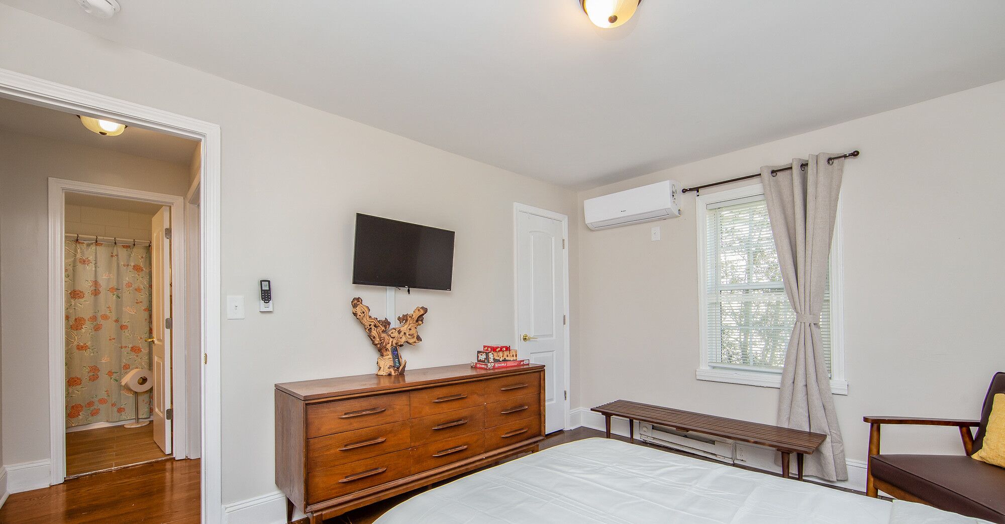 A bedroom with a bed, wooden dresser, TV, air conditioning unit, window with curtains, and a bathroom with floral shower curtain.