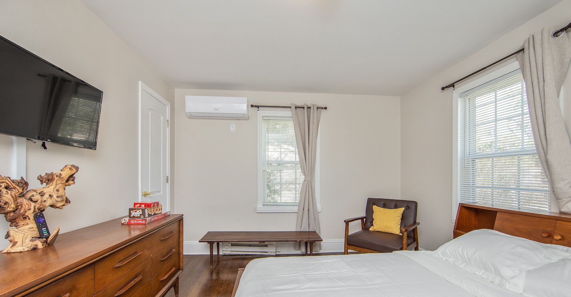 A bedroom with a bed, a wooden dresser, a chair, a bench, a television, and an air conditioning unit.