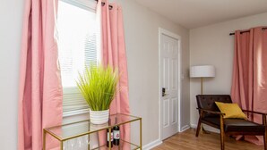 A room with pink curtains, a glass table with wine bottles, a sofa with a yellow pillow, and a lamp. - The Ramsey Rose Residence (Fayetteville)