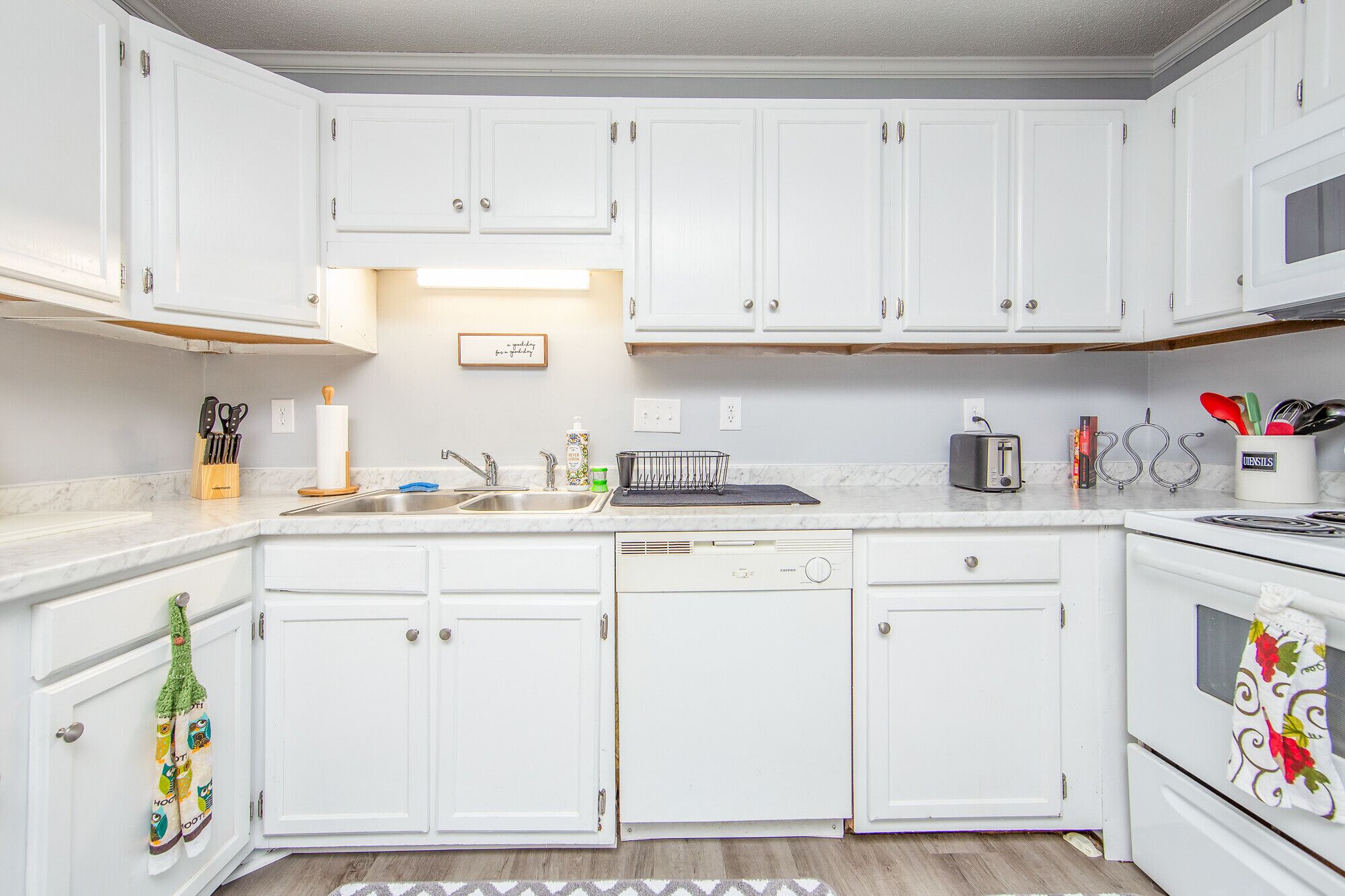 A kitchen with white cabinets, a dishwasher, a toaster, and a microwave. There is a wooden cutting board and a knife block on the counter.