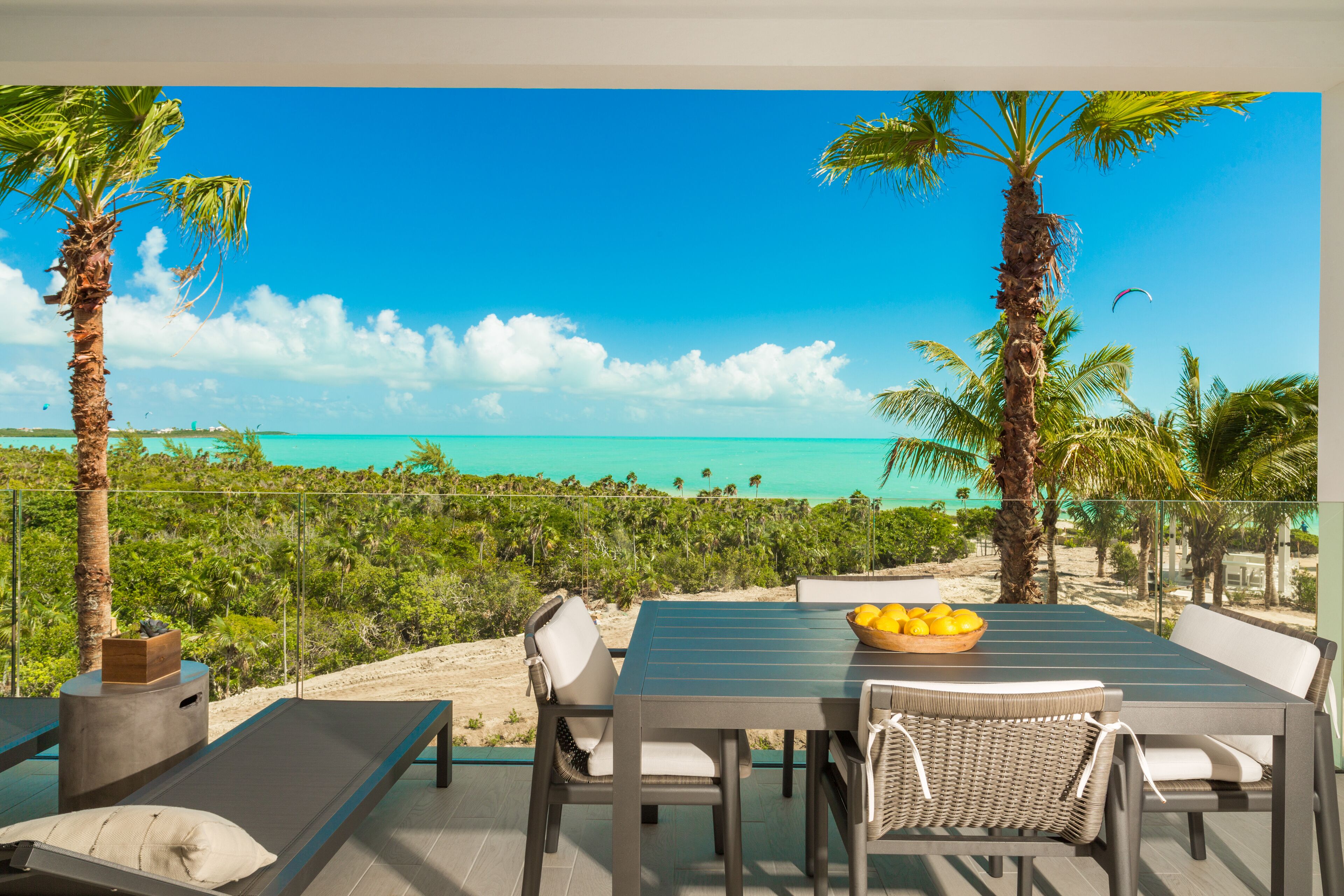 A balcony with a table and chairs overlooking a beach with palm trees and a clear sky.