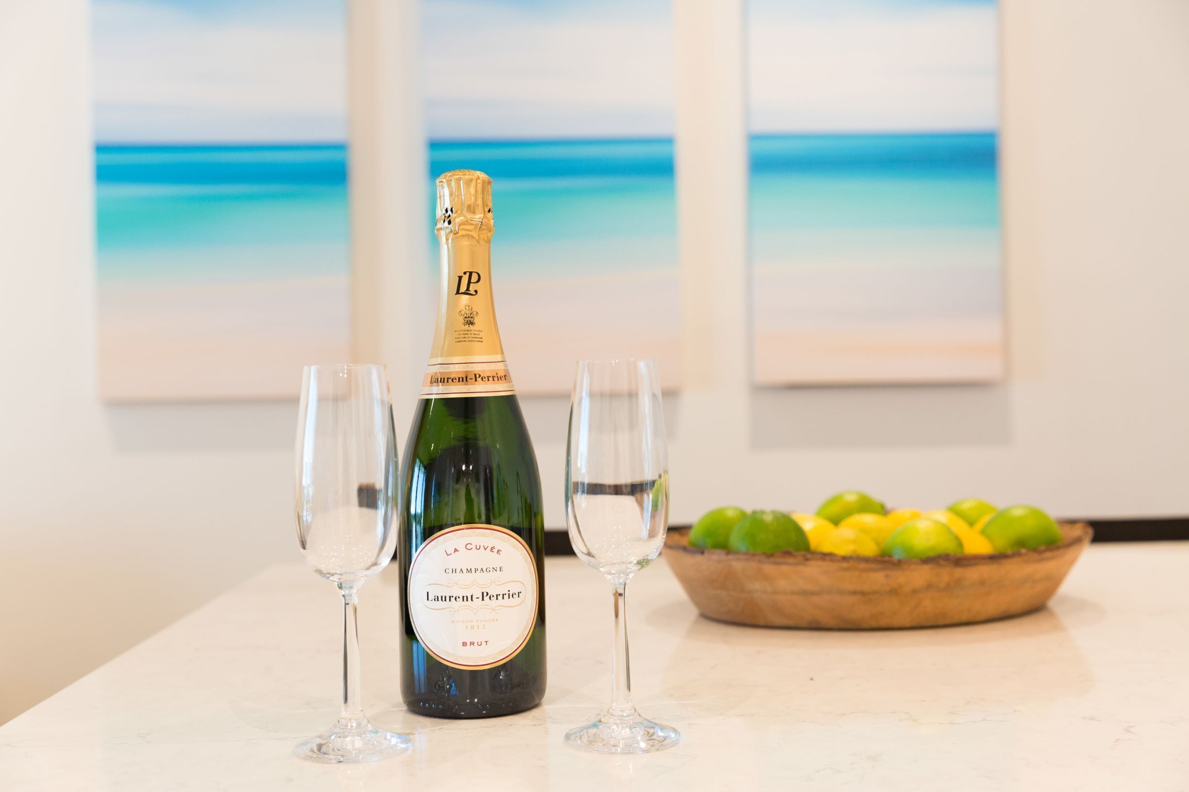 A bottle of champagne with two glasses and a bowl of fruit on a table.