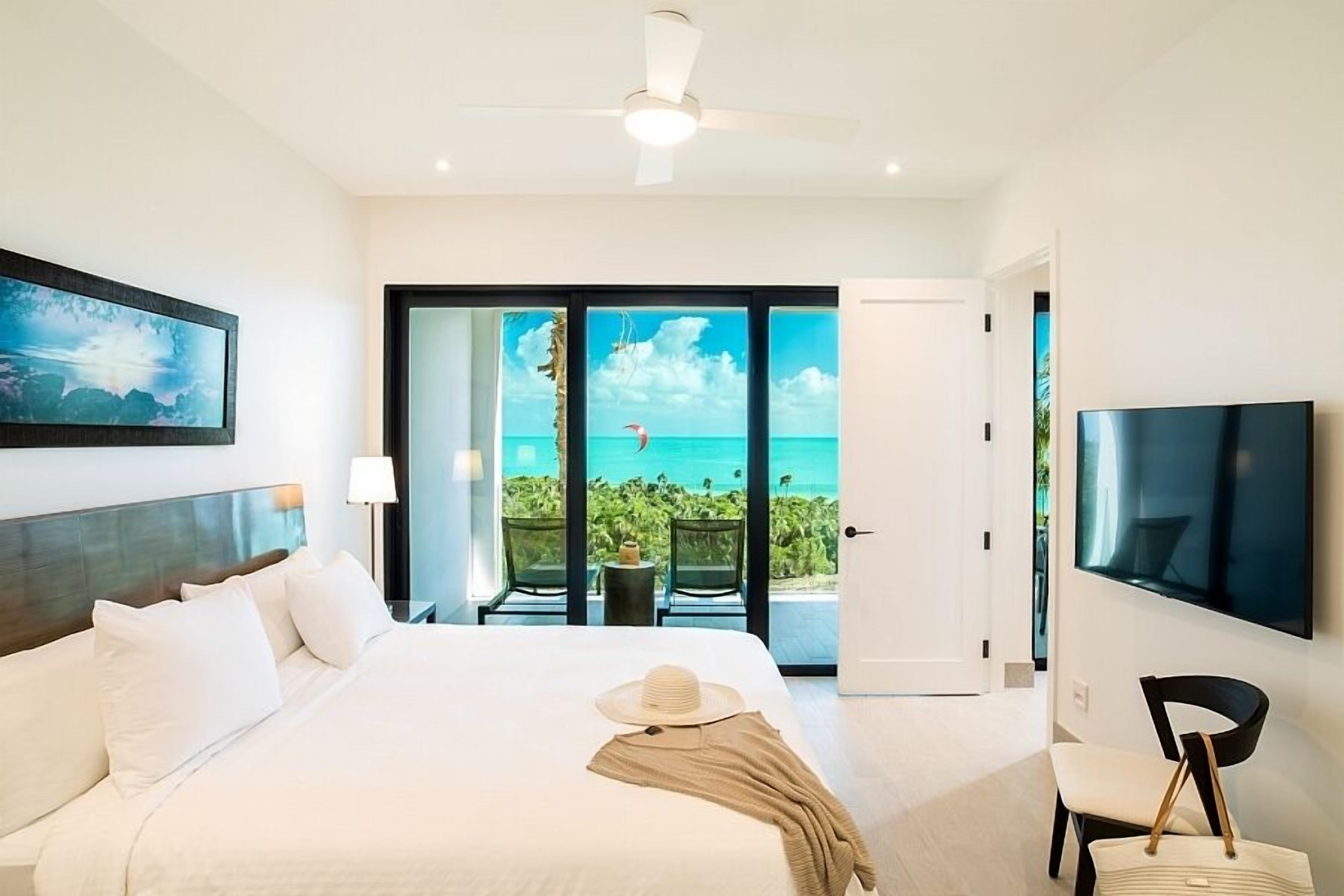 A hotel room with a bed, a TV, a balcony with a view of the ocean, and a ceiling fan.