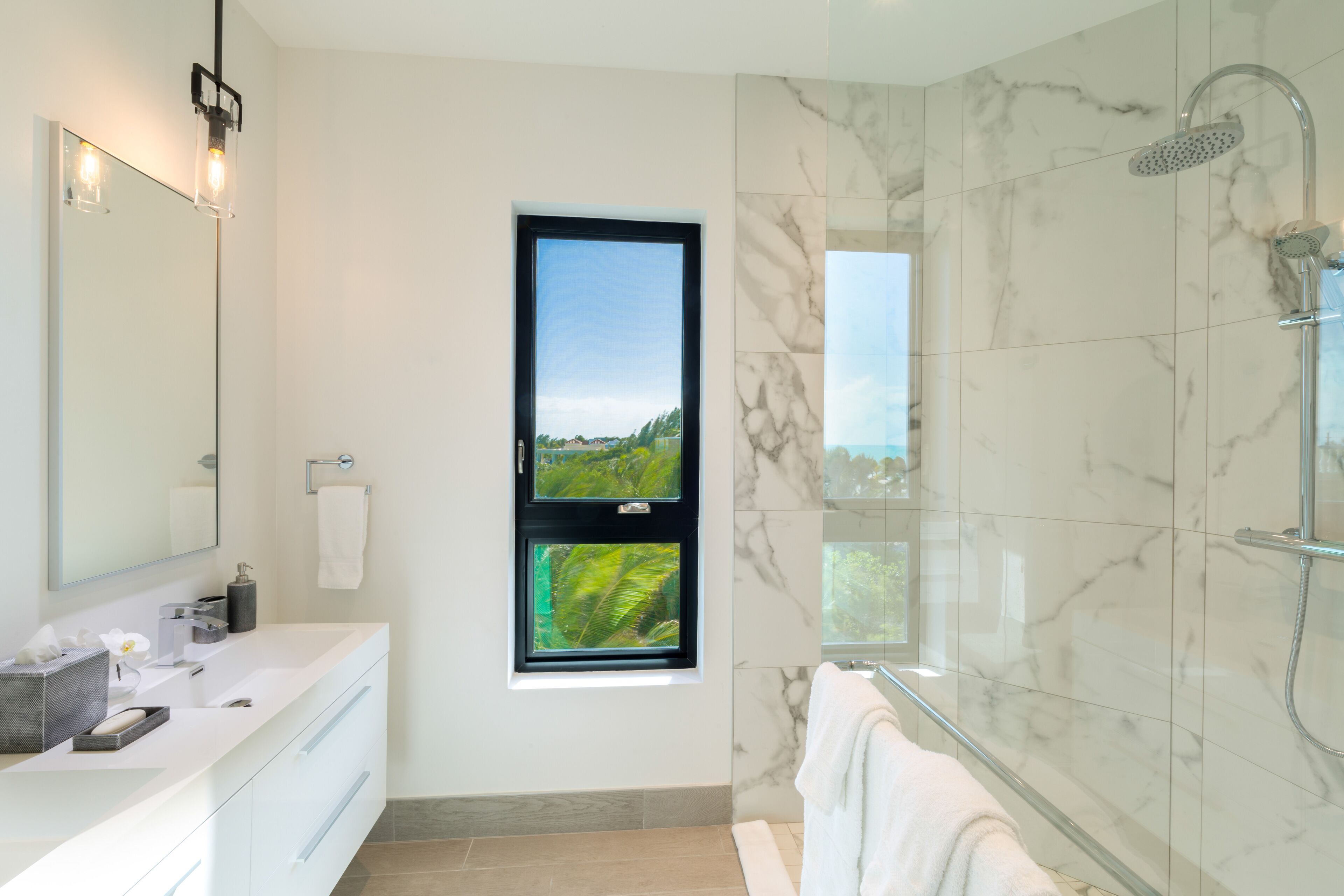 A modern bathroom with a large mirror, a window with a view of greenery, a shower area with a glass door, and a white vanity with a sink.