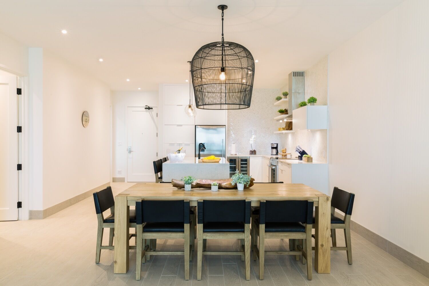 Un espace repas moderne avec une table et des chaises en bois, un luminaire suspendu et une cuisine en arrière-plan.