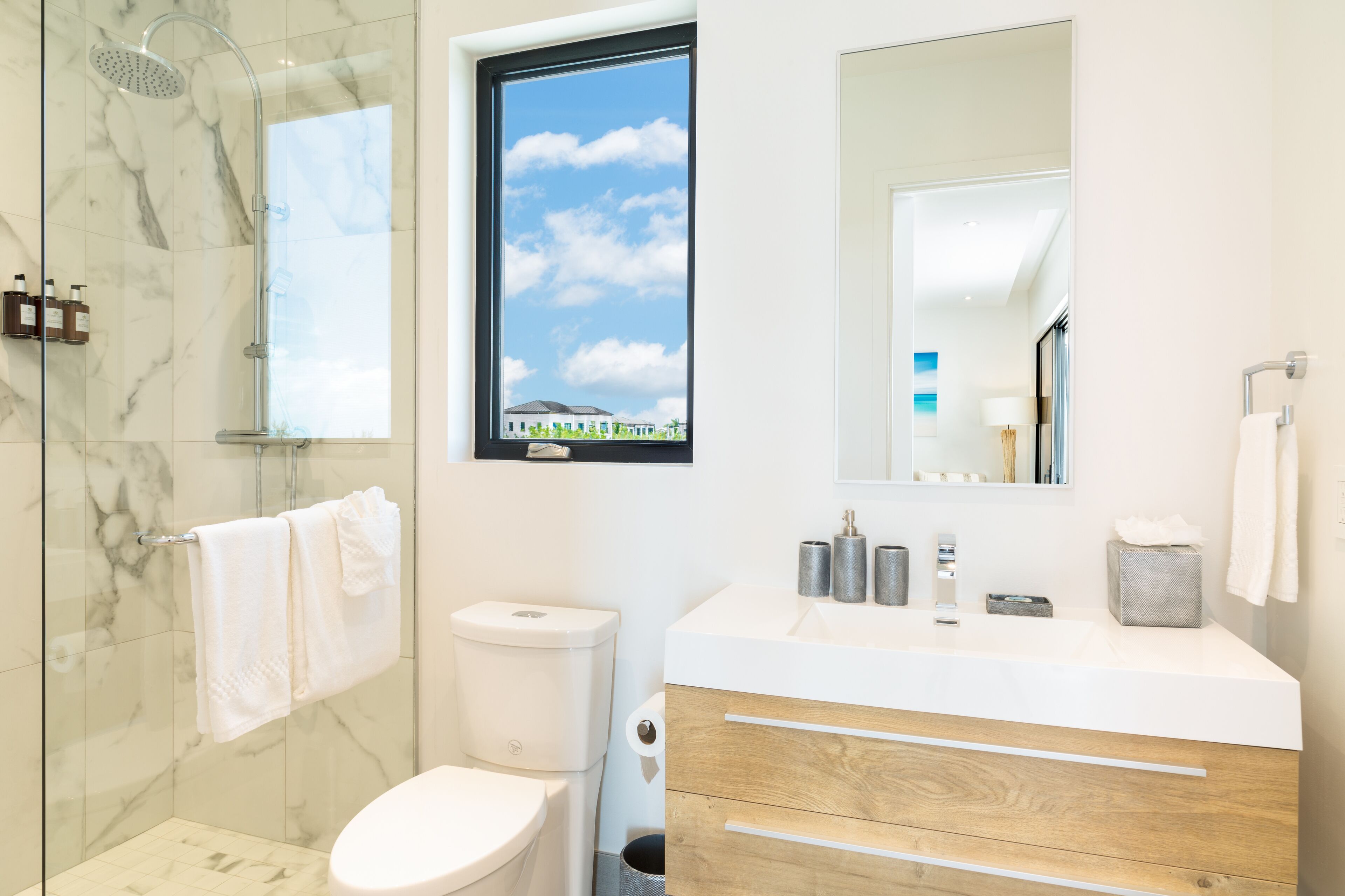 A modern bathroom with a glass-enclosed shower, a toilet, a sink with a mirror above it, and a window providing a view of the sky.