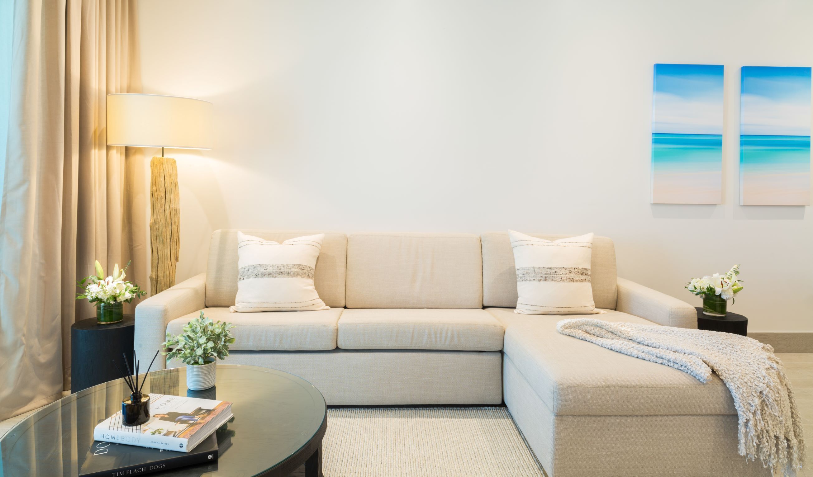 A modern living room with a beige sofa, a round coffee table, and a lamp. There are two side tables with decorative items and a book on the coffee table.