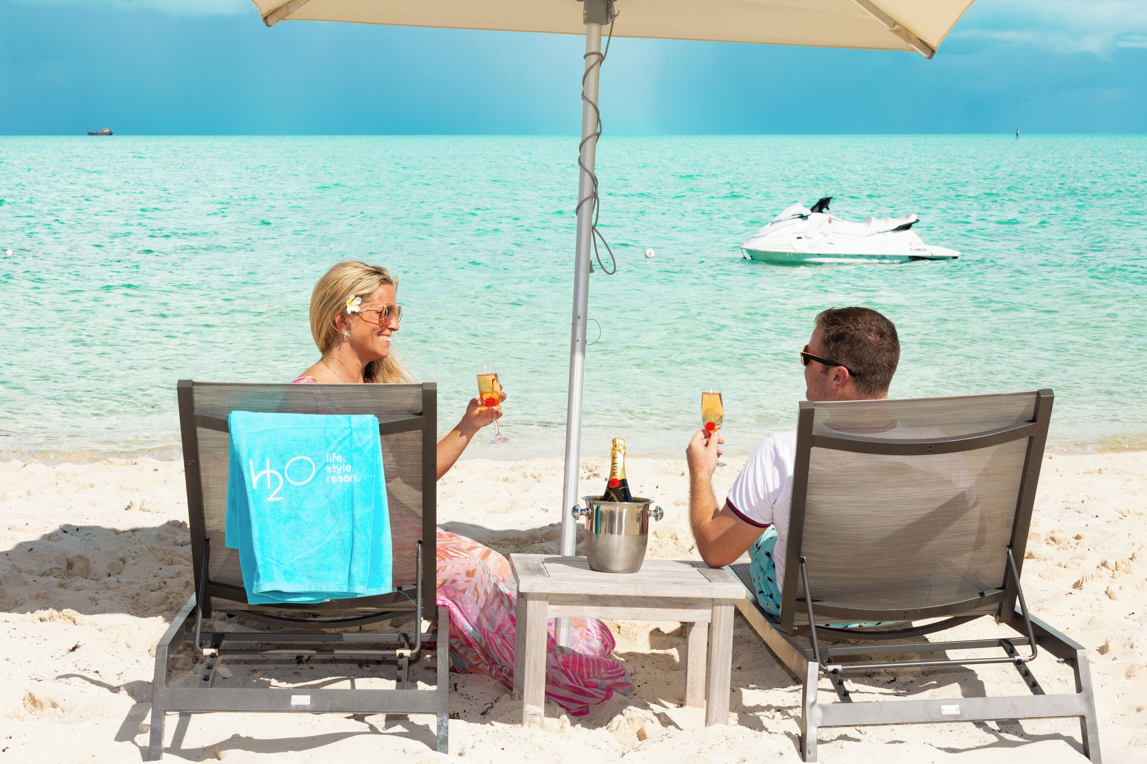 Two people sitting at a table with a bucket of champagne on a beach.