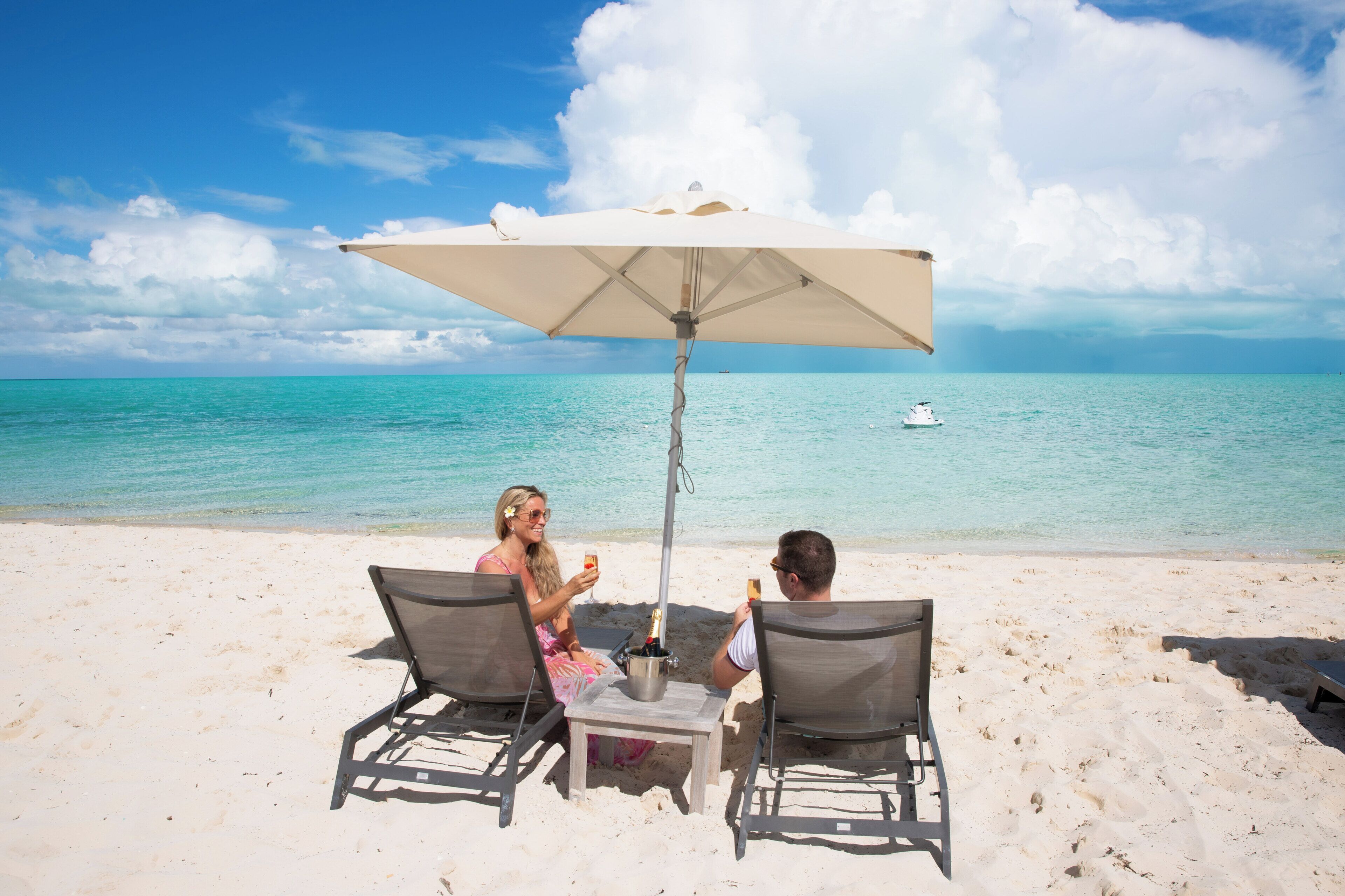 Two people sitting at a table with a beach umbrella on a sandy beach with a boat in the background.