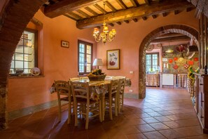 A dining area with a wooden table and chairs, a chandelier, and a kitchen visible through an archway. - Villa Montelopio (Peccioli)