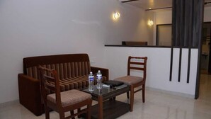 A room with a brown sofa, two wooden chairs, a glass coffee table, and a small kitchen area with a counter and cabinets. - The Byke Sai Cottage, Mahabaleshwar (Satara)