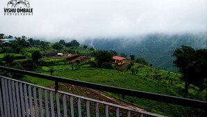 A lush green landscape with terraced fields and a few buildings, including a red-roofed structure. - The Byke Sai Cottage, Mahabaleshwar (Satara)