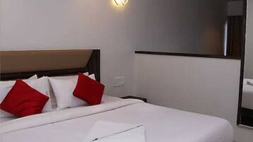 Habitación de hotel con cama, ropa blanca, almohadas rojas, mesita de noche y una lámpara de pared.