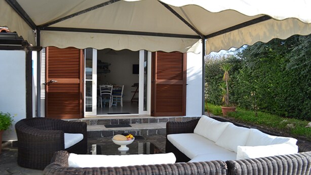 Un porche exterior cubierto con mobiliario de mimbre, una mesa con un cuenco de fruta y vistas a un jardín.