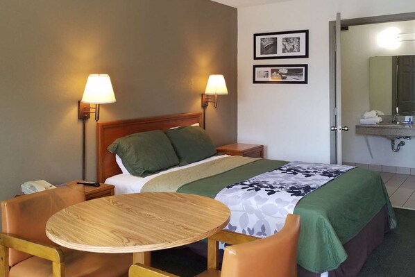 Habitación de hotel con cama, dos lámparas de noche, una mesa redonda de madera, dos sillas, baño con lavabo y espejo, y dos cuadros en la pared.