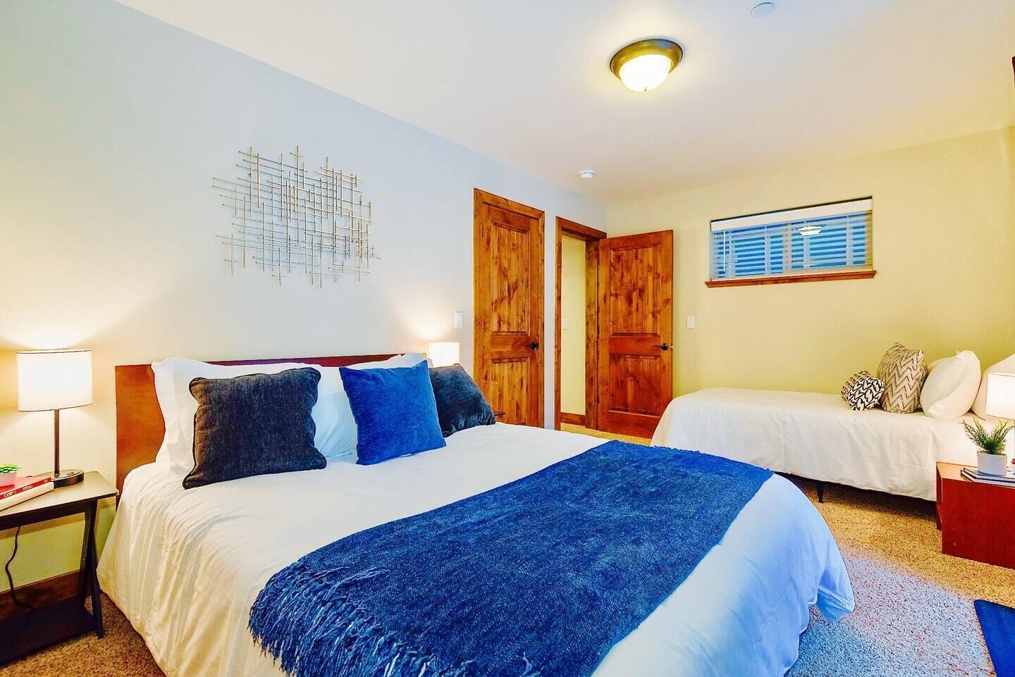 A hotel room with a large bed, a smaller bed, wooden doors, a window with blinds, and a wall-mounted artwork.