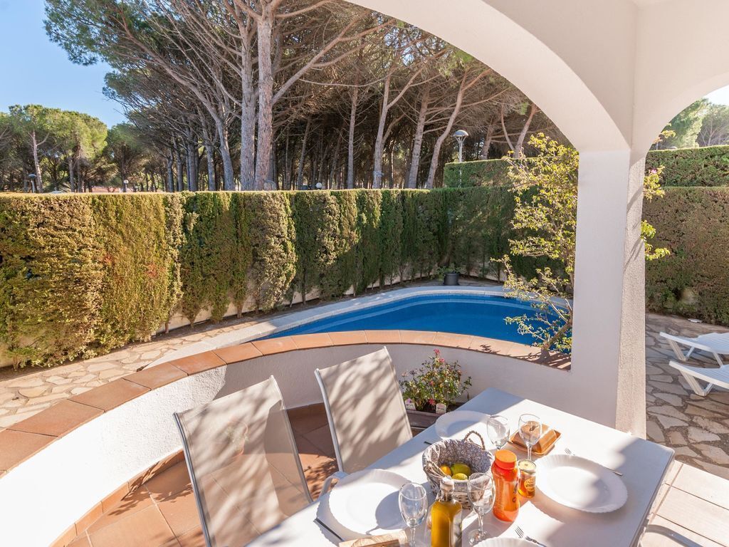 Un área de comedor junto a la piscina con una mesa dispuesta para dos, rodeada por un arco blanco, con vistas a una piscina y un jardín bordeado de setos.