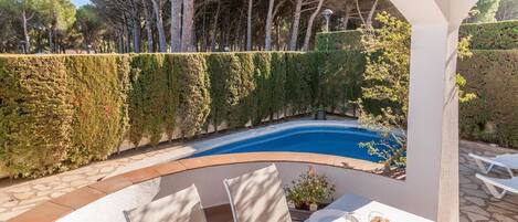Un área de comedor junto a la piscina con una mesa dispuesta para dos, rodeada por un arco blanco, con vistas a una piscina y un jardín bordeado de setos.