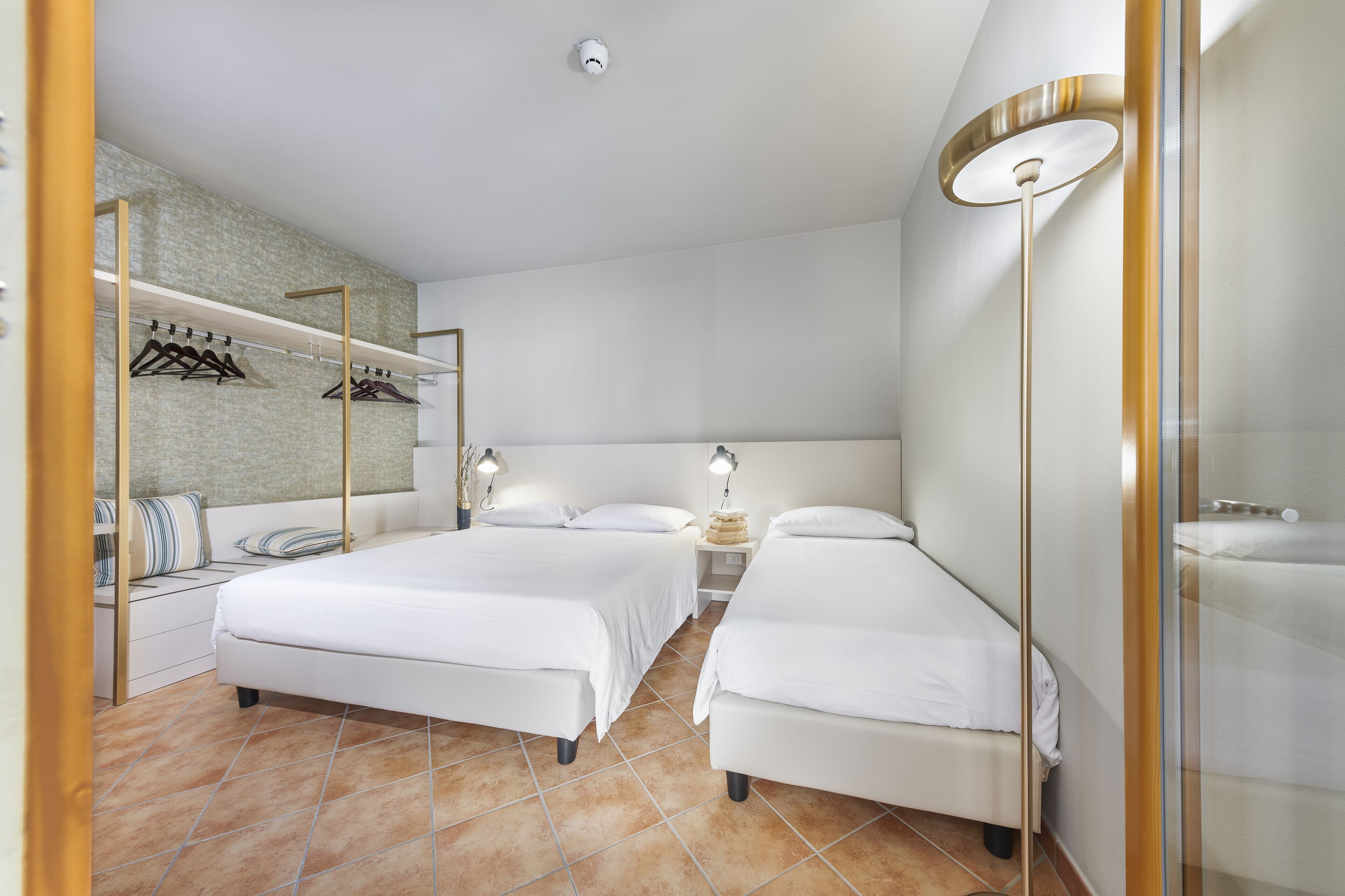 A modern hotel room with two beds, a seating area with a bench and pillows, a small bedside table with a lamp, and a wall-mounted shelf with hangers.