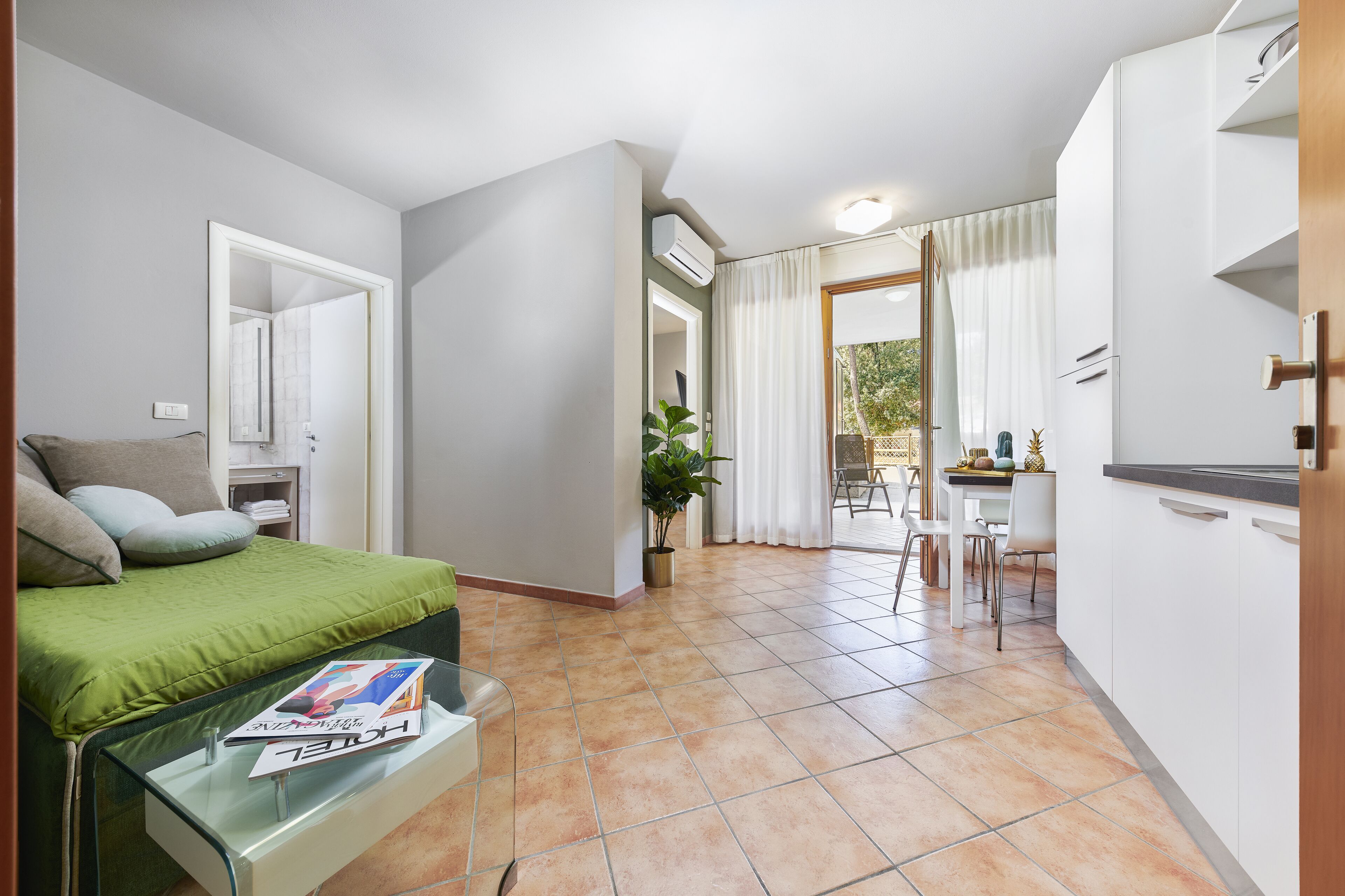 A compact hotel room with a bed, a glass table with a magazine, a dining area with a table and chairs, and a kitchenette.