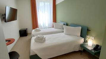 A hotel room with two beds, a TV mounted on the wall, a desk with a chair, and a nightstand with a lamp.