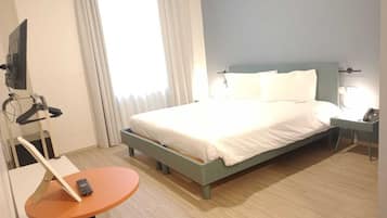 Ett hotellrum med en säng, ett skrivbord med en stol, en tv som är monterad på väggen och ett fönster med gardiner.