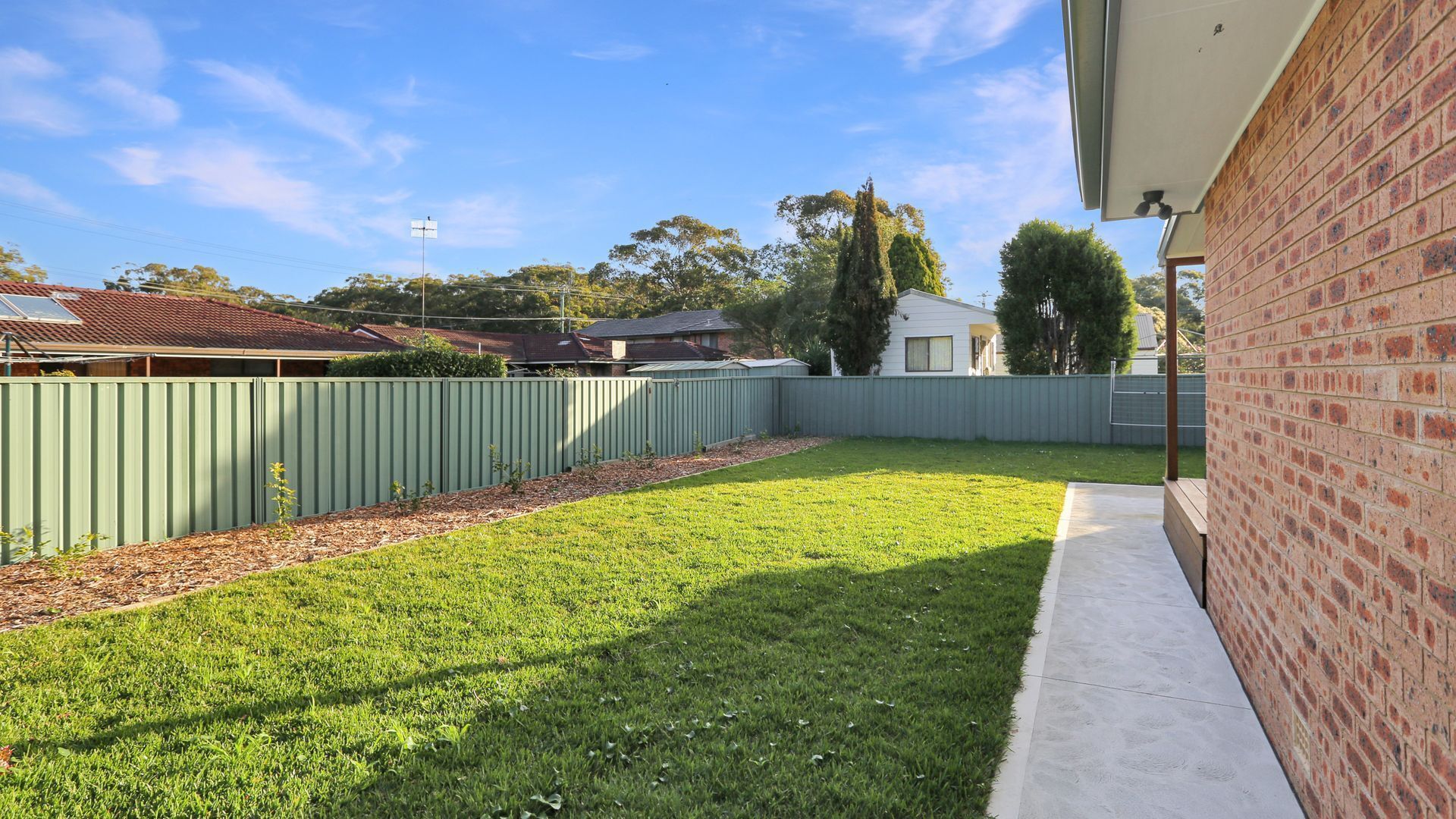 A backyard with a green fence, a grassy lawn, and a brick wall.