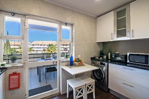 A compact kitchen with a washing machine, a microwave, and a small table with a basket of fruit. - "Vilamoura Boutique" Apartment with pool, Wi-Fi, air conditioning and balcony (Quarteira)