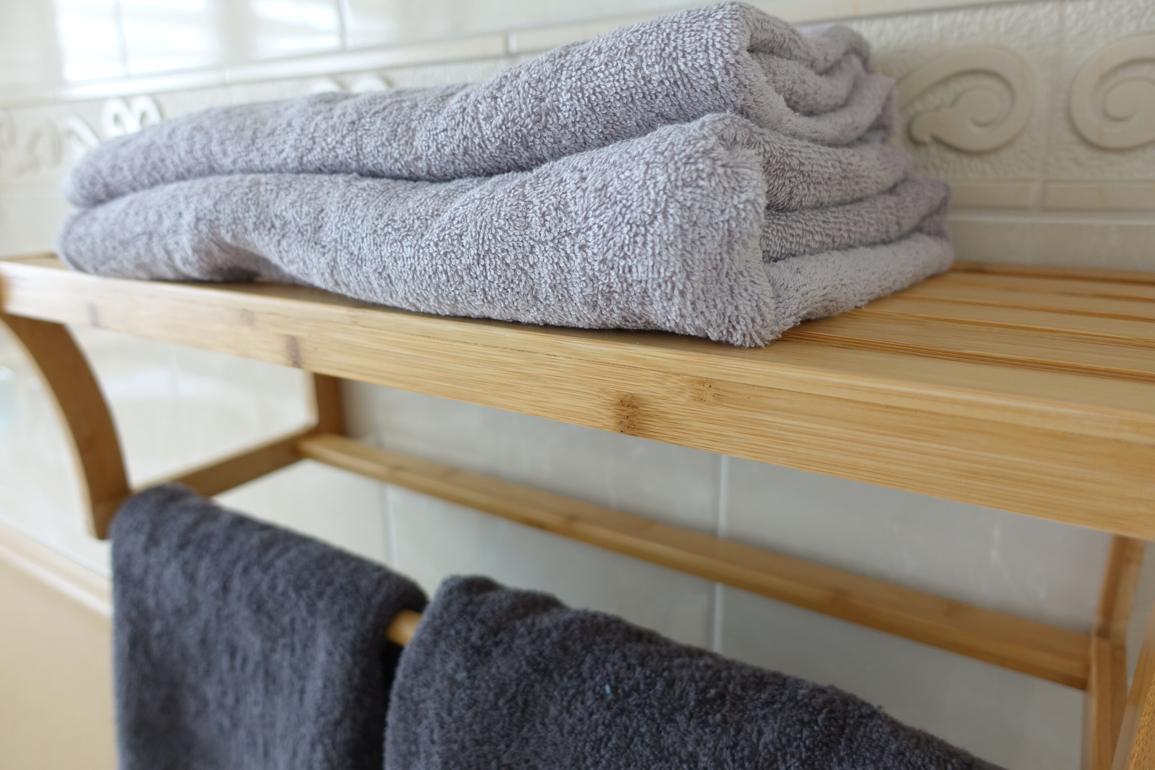 A stack of folded towels on a wooden shelf in a bathroom with white tiled walls.