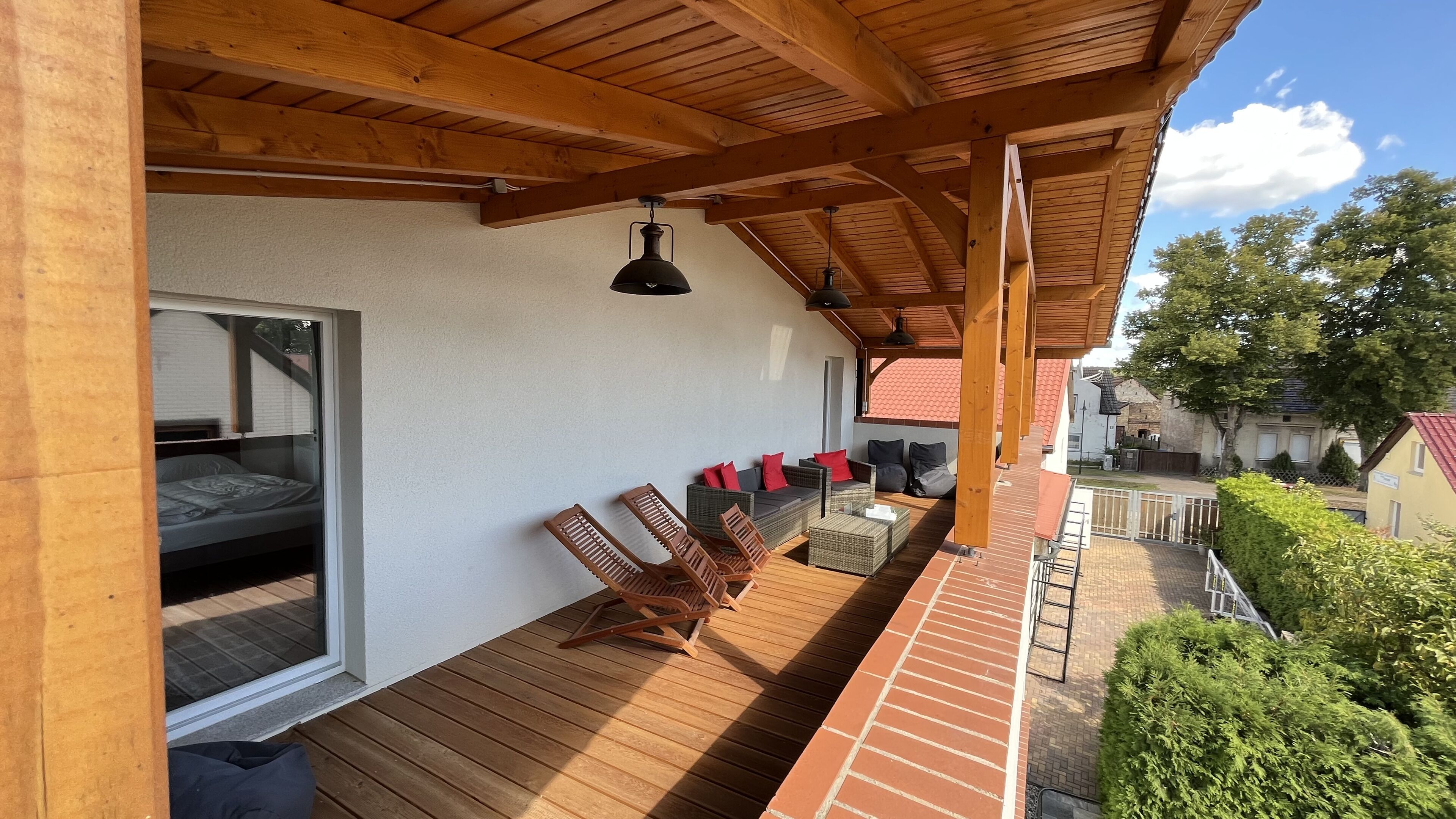 A covered outdoor seating area with wooden chairs and tables, a window with a bed inside, and a view of the surrounding neighborhood.