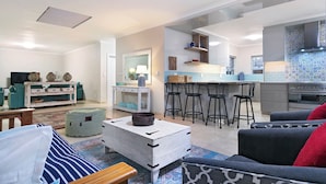 A modern living room with a sofa, a coffee table, and a TV. A kitchen with bar stools and a tiled backsplash is visible through an open doorway. - Melkbos Aan See (Cape Town)