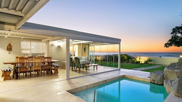 A modern outdoor pool area with a glass enclosure, a dining set, and a view of the ocean at sunset. - Melkbos Aan See (Cape Town)