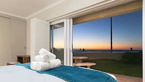 A bedroom with a bed, a blue blanket, and white towels. There is a sliding glass door leading to a balcony with a view of the beach and ocean at sunset. - Melkbos Aan See (Cape Town)